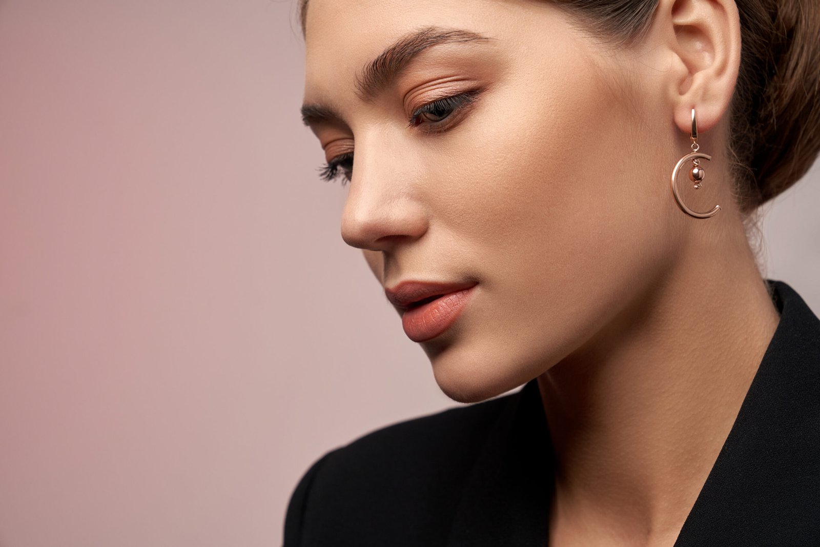Close up portrait of young beautiful brunette female model presenting golden earrings. Side view of woman in formal suit posing, isolated on studio background. Concept of jewelry.